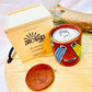 100% natural soy wax scented candle in a terracotta jar, hand painted with Indian classical musical instruments motif, and covered with seed paper candle dust cover is placed on a wooden tray. A terracotta candle snuffer is placed near the scented candle with a wooden candle box and honeycomb paper in the background.