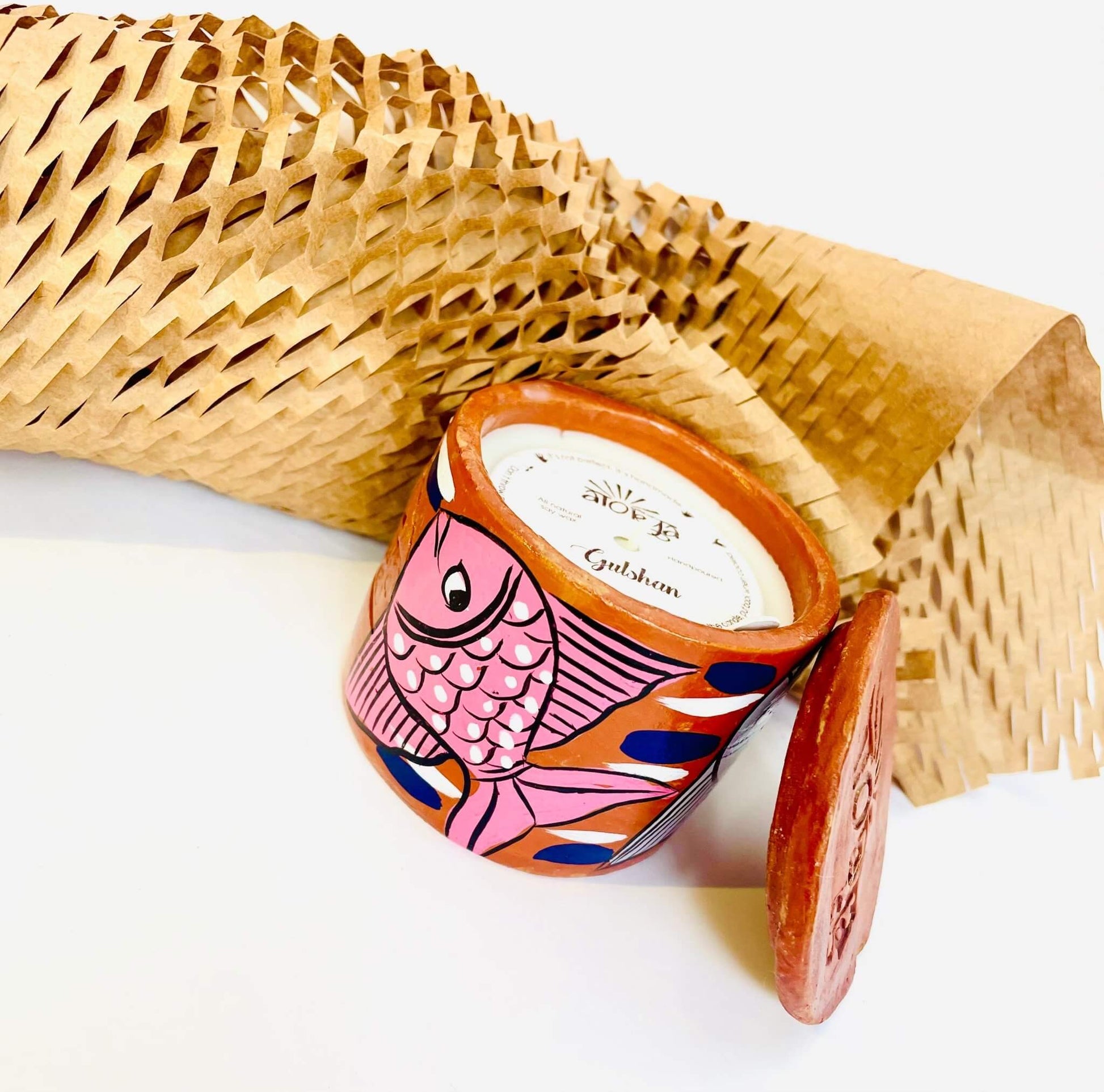 Scented candle in a terracotta jar painted with pink fish. The candle is protected with seed paper while candle snuffer and honeycomb paper are placed around it.