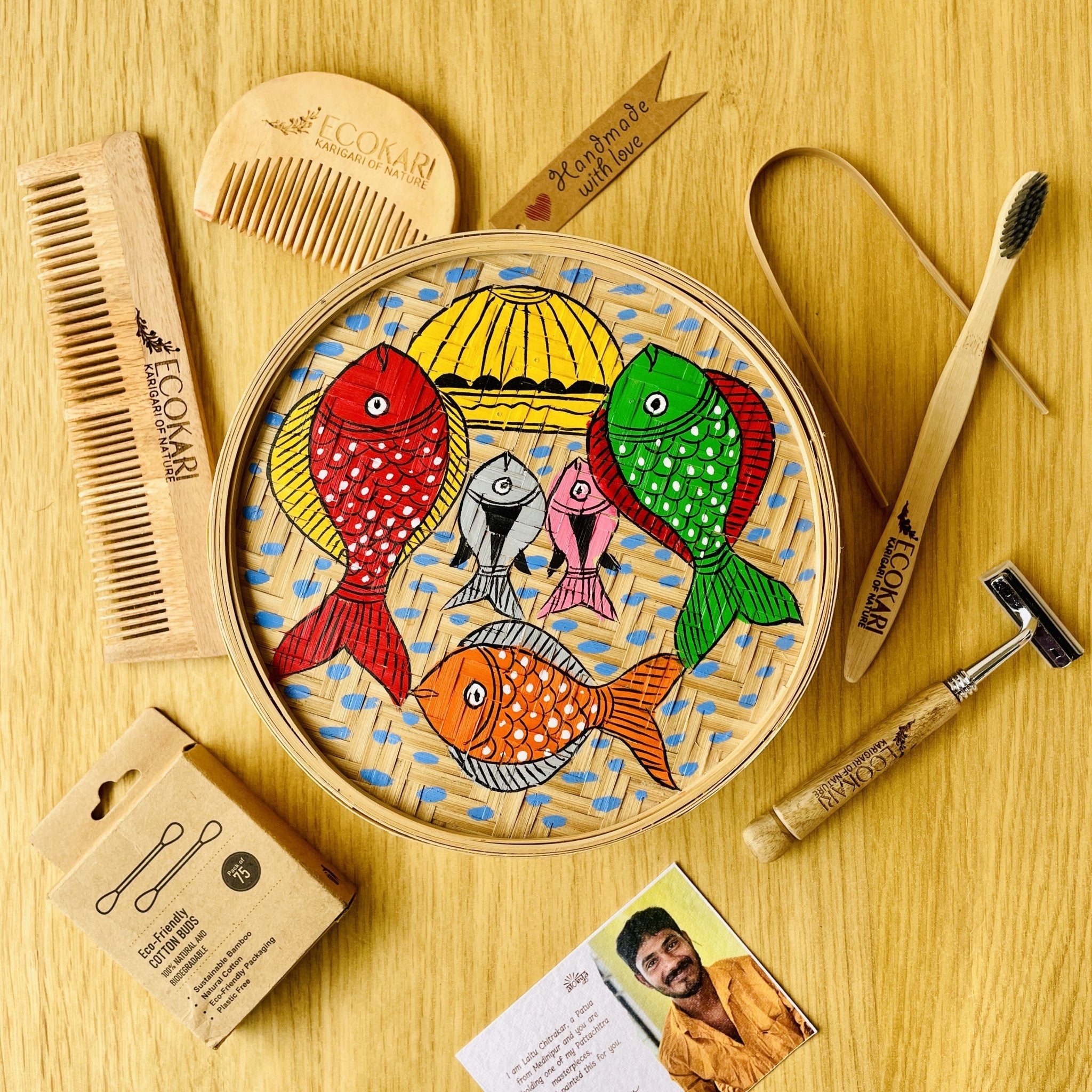 Men's neem wood beard comb, unisex neem wooden comb, cotton bamboo earbuds, tongue cleaner, bamboo toothbrush, bamboo wooden shaving razor displayed around a bamboo box hand painted with fish paintings in traditional Pattachitra art form style.