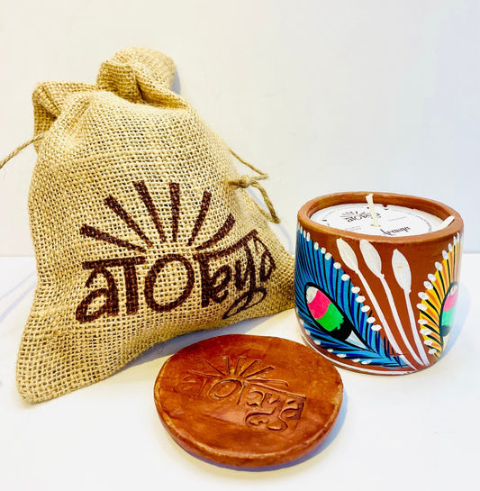 100% natural soy wax scented candle in a terracotta jar, hand-painted with yellow and blue feathers motif is covered with seed paper dust cover and a jute bag and terracotta clay candle snuffer is placed near the aromatherapy scented candle.
