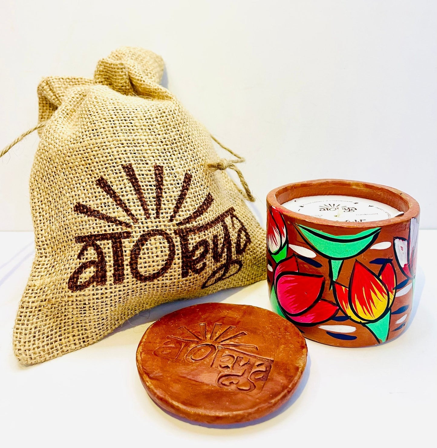 100% natural soy wax scented candle in a terracotta jar, hand-painted with one red lotus and one orange-yellow lotus surrounded by green leaves motif is covered with seed paper dust cover and a jute bag and terracotta clay candle snuffer is placed near the aromatherapy scented candle.