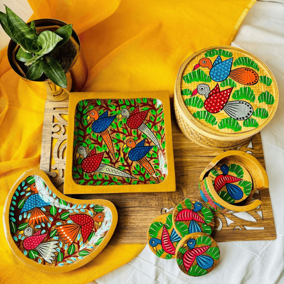 pure mango wooden platters, set of 6 round wooden coasters and handwoven bamboo utility box, all hand painted with bird motifs in traditional pattachitra art are displayed on a rectangle wooden board against a yellow backdrop beside a golden flower pot