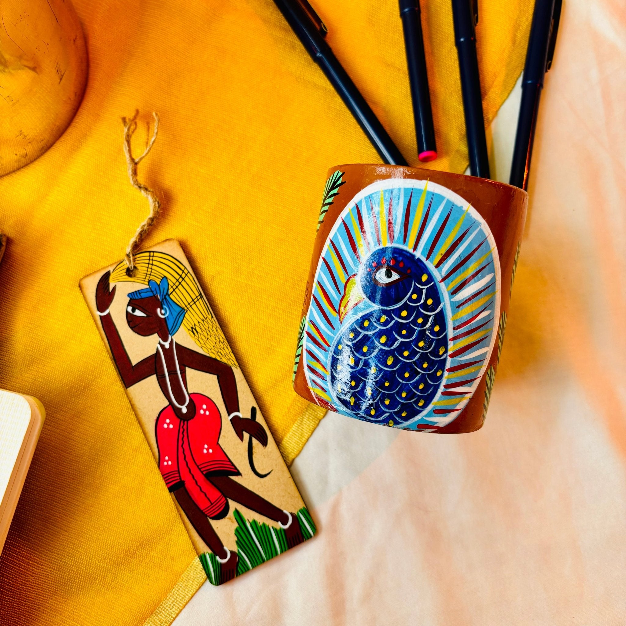 Terracotta clay pen and pencil stand/holder hand painted with peacock painting and a wooden bookmark hand painted with tribal painting are displayed near four black pens against a yellow backdrop