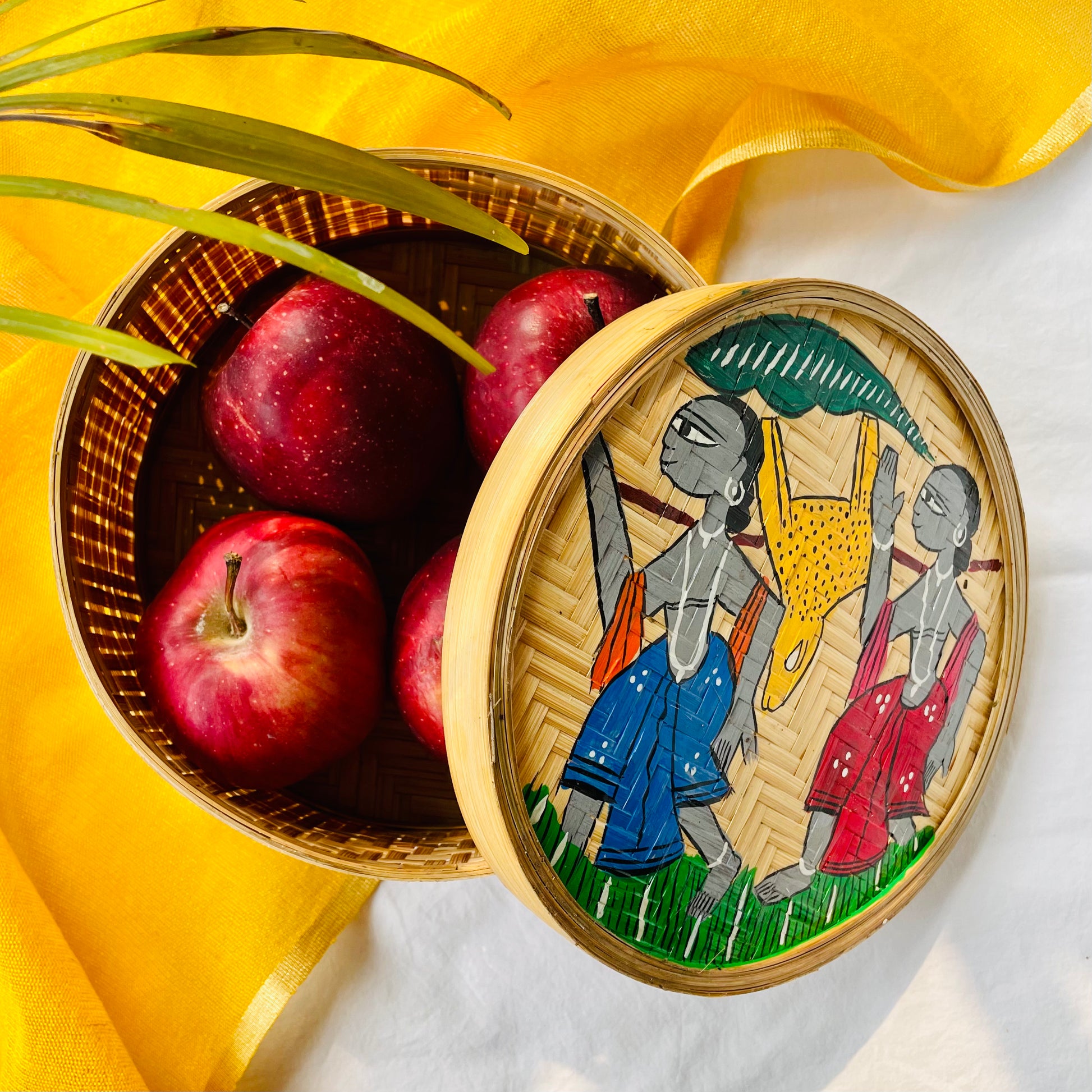 A handwoven bamboo box hand painted with a motif of tribal characters and filled with four apples is displayed against a yellow and white background