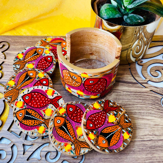 an empty coaster holder placed in front of a plant pot along with 6 round wooden coasters, all hand-painted with red and orange fishes having pink and yellow fins motifs.