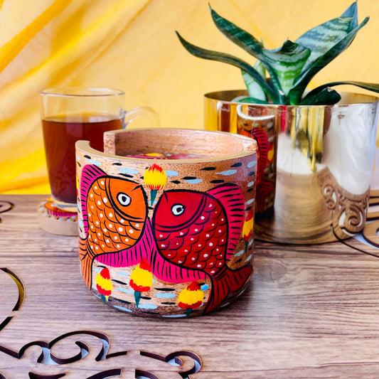 pure mango wood round wooden coaster holder with red and orange fishes having pink and yellow fins painted on it is being displayed with a warm cup of black tea and a plant pot in the background.