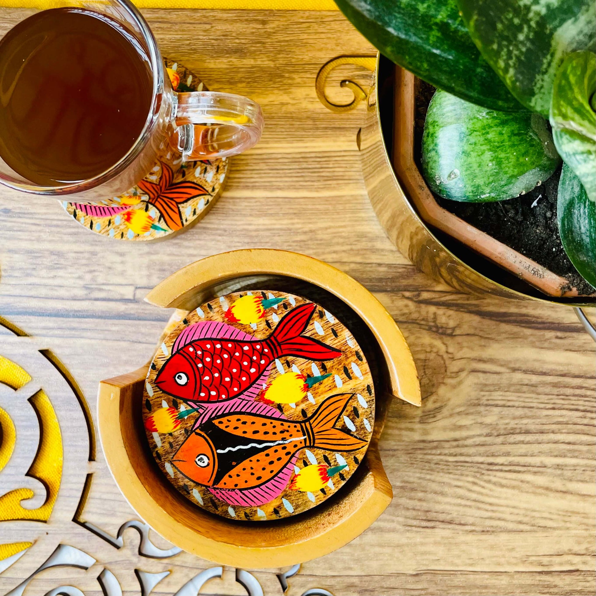 red and orange fishes having pink and yellow fins print round wood coasters placed in a round wooden coasters holder near a flower pot and tea cup placed on a round wooden coaster.