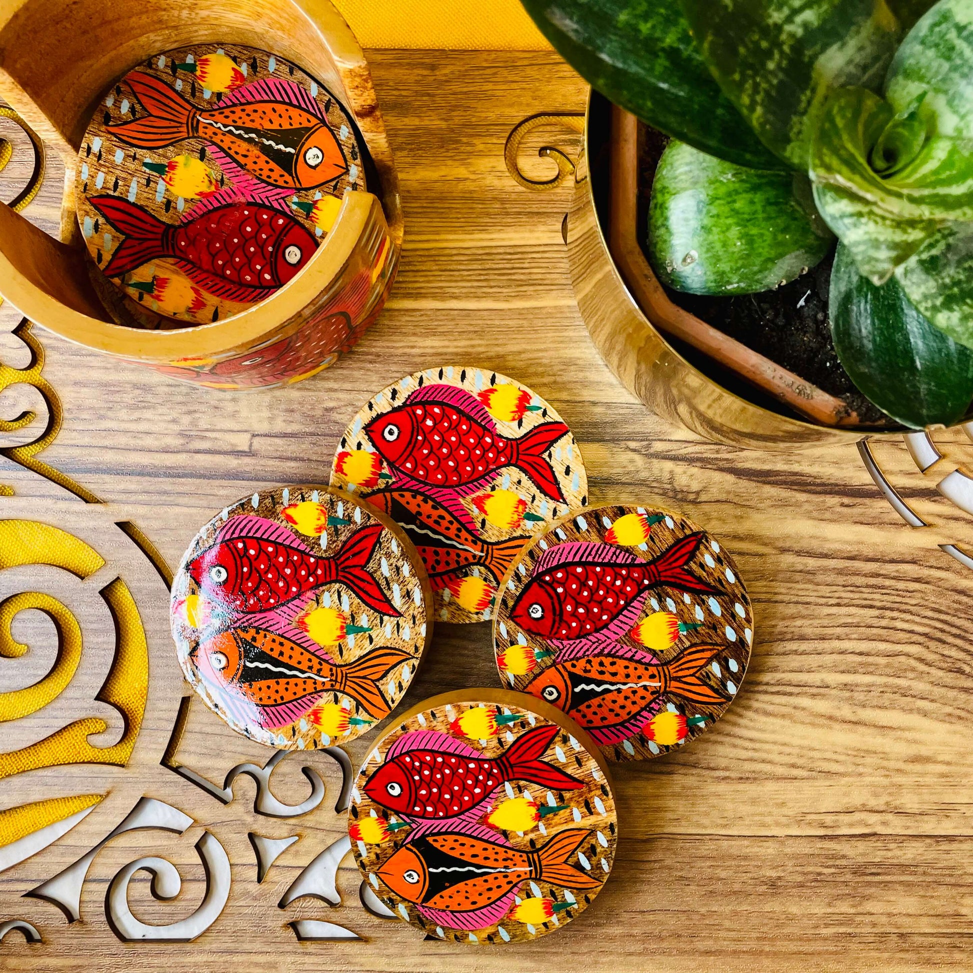 One pure mango wood round wooden coaster is placed in a wood coaster holder while 4 round wooden coasters hand-painted with red and orange fishes having pink and yellow fins are displayed near a flower pot.