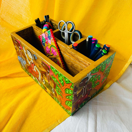 An aerial view of three pens, two scissors, six coloured pens and a bookmark placed in a 9”x5”x3” pure mango wood table organiser or wood cutlery holder wooden tissue holder with 3 small and one large compartment, handcrafted by rural artisans and painted with a scene of tribal celebration and trees
