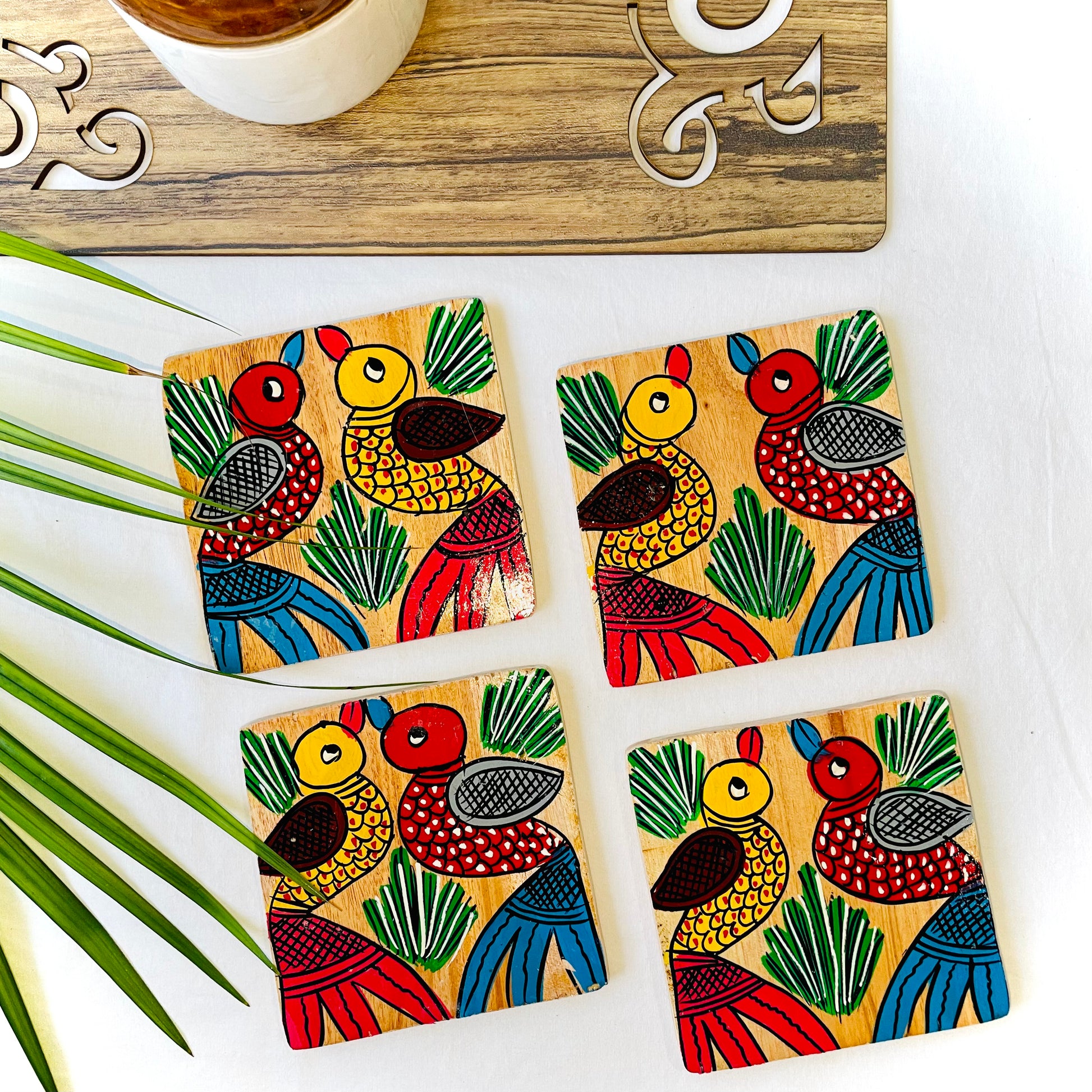 Four pure mango wood square wooden coasters, with a painting of one yellow bird with red feathers and a beak and one red with blue feathers and a beak along with some leaf in each wood coaster