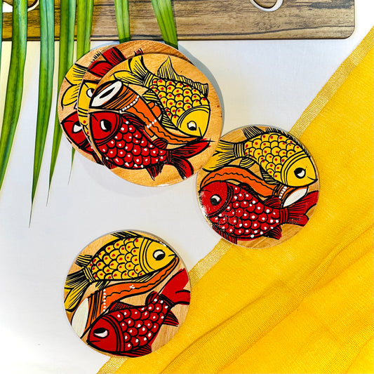 Three pure mango wood round wooden coasters, hand painted with one yellow fish and one redfish along with an Indian classical musical instrument in each wood coaster are placed against a yellow backdrop and leaves in the background