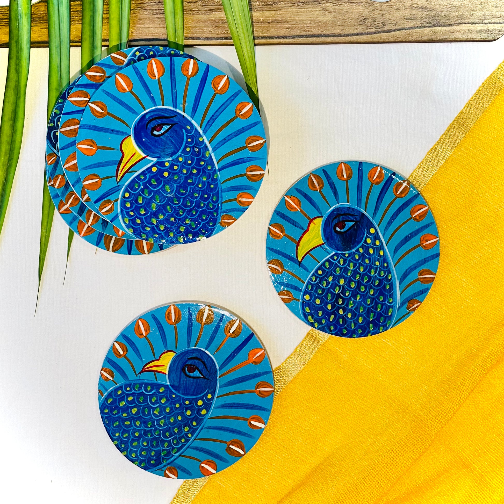 Three pure mango wood round wooden coasters, hand painted with peacocks with blue bodies, red feathers and yellow beaks are placed against a yellow backdrop and leaves in the background