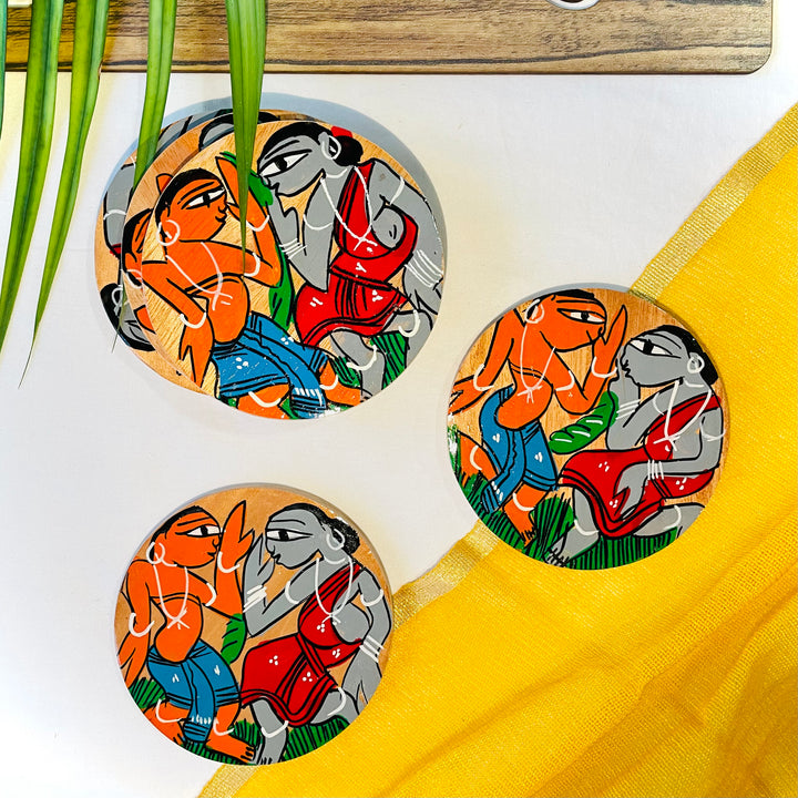 Three pure mango wood round wooden coasters, hand painted with tribal characters dancing placed against a yellow backdrop and leaves in the background