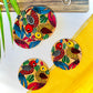 Three pure mango wood round wooden coasters, hand painted with one yellow bird with red feathers and a beak and one red with blue feathers and a beak along with some leaf in each wood coaster are placed against a yellow backdrop and leaves in the background