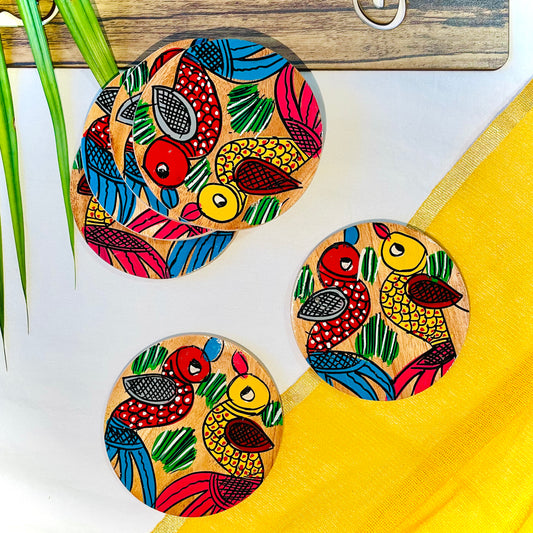 Three pure mango wood round wooden coasters, hand painted with one yellow bird with red feathers and a beak and one red with blue feathers and a beak along with some leaf in each wood coaster are placed against a yellow backdrop and leaves in the background