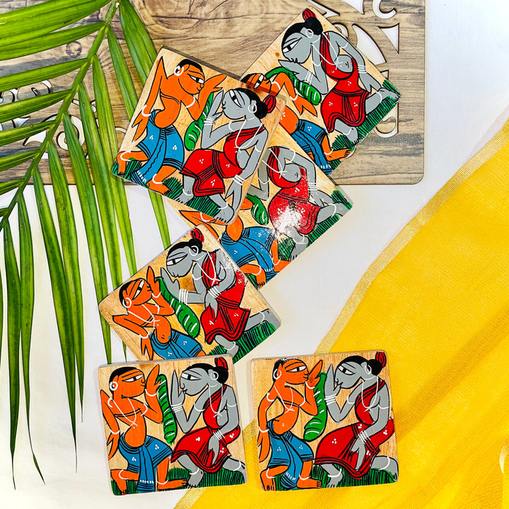 Six pure mango wood square wood coasters hand painted with tribal characters dancing are placed on a wood tray with leaves in the background