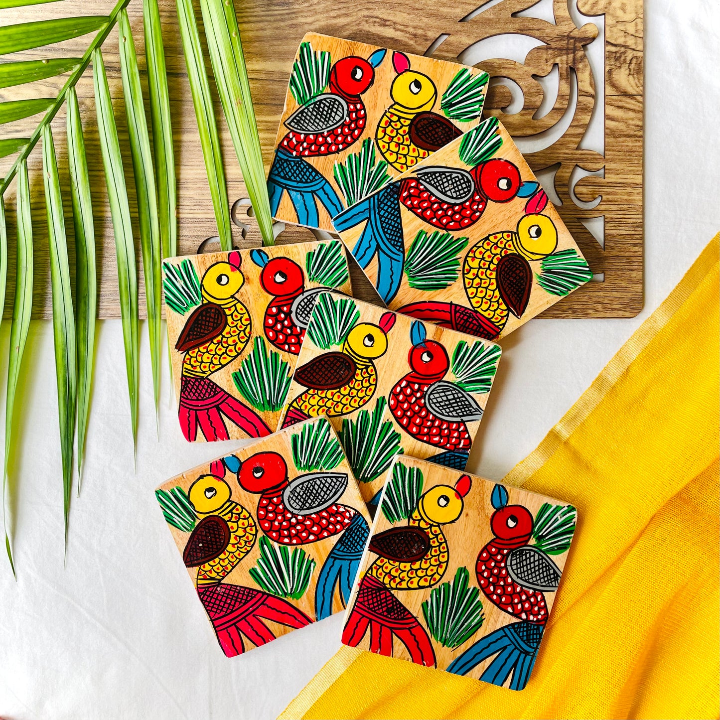 Six pure mango wood square wood coasters painted with a motif of one red-coloured bird with blue wings and beak and one yellow-coloured bird with red Wings and beak on each coaster are placed on a brown wood board with leaves in the background