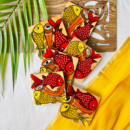 Six pure mango wood square wood coasters painted with one yellow fish and one redfish along with an Indian classical musical instrument in each wood coaster are placed on a wood tray with leaves in the background