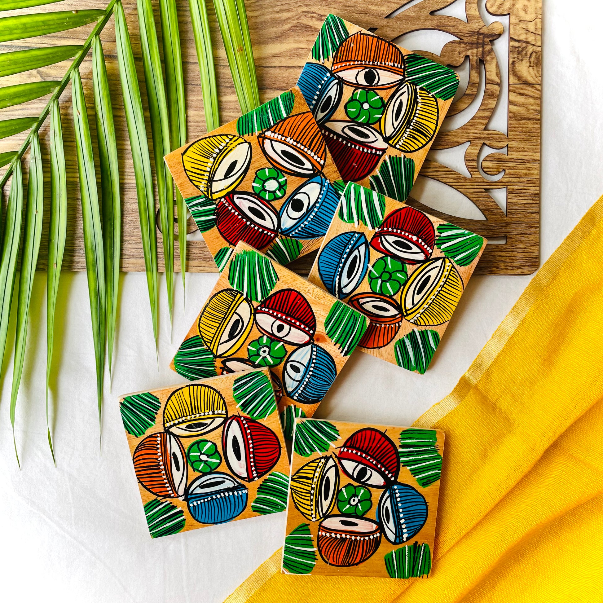 Six pure mango wood square wood coasters painted with four tablas, a type of Indian classical musical instrument in each wood coaster are placed on a wood tray with leaves in the background