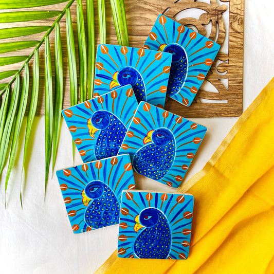 Six pure mango wood sqaure wood coasters painted with peacocks having blue bodies, red feathers and yellow beaks are placed on a wood tray with leaves in the background