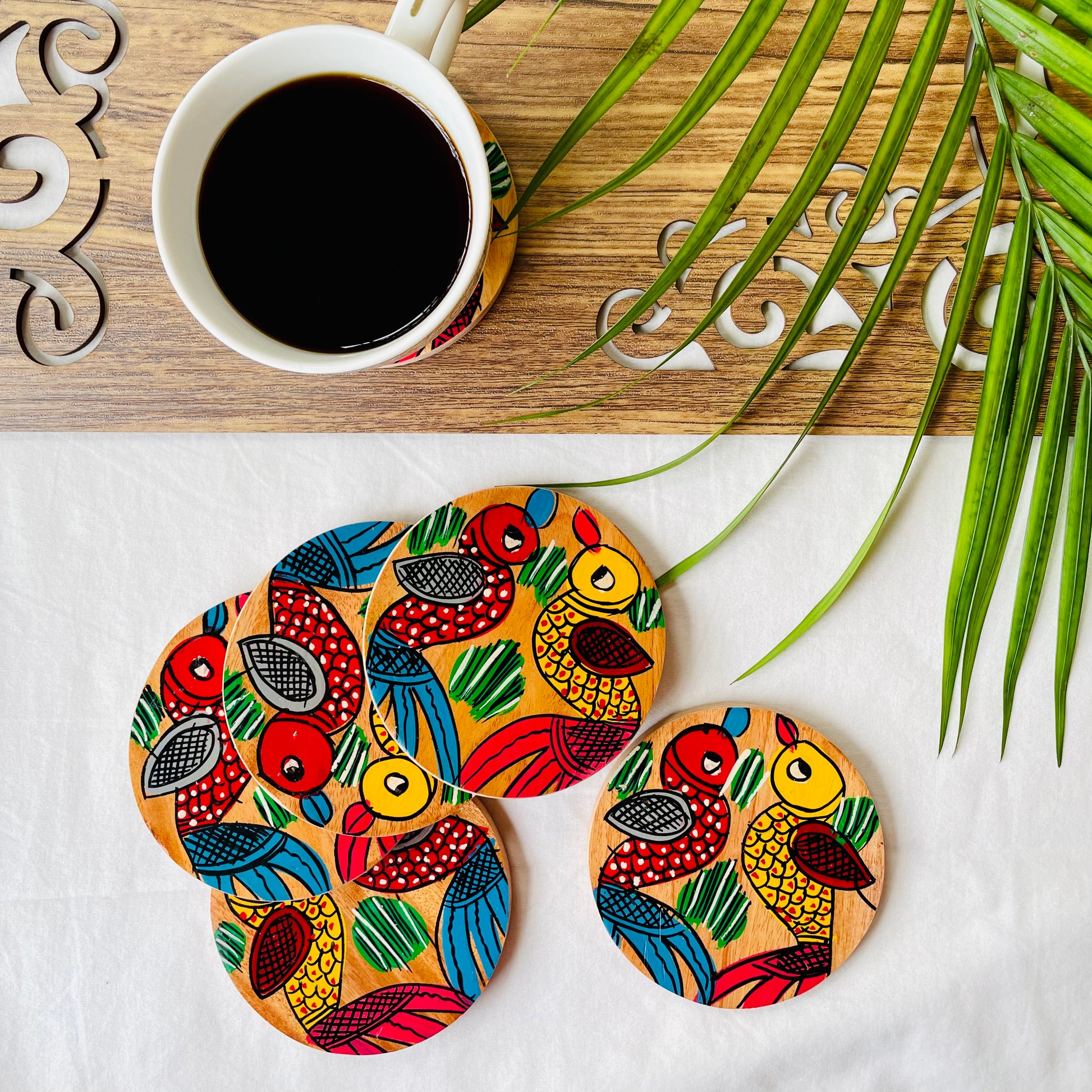 Five pure mango wood round wood coasters painted with one yellow bird with red feathers and a beak and one red with blue feathers and a beak along with some leaf in each wood coaster are placed near a teacup-filled with black tea with leaves in the background