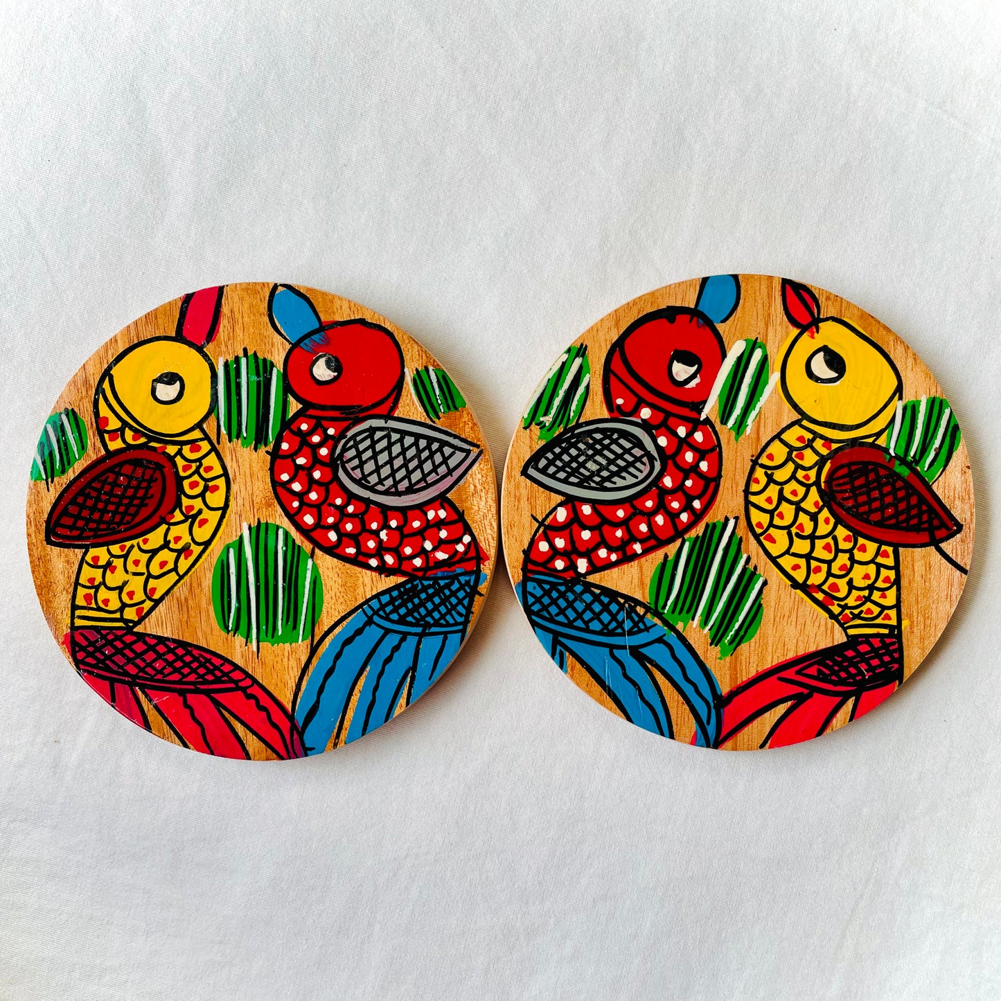 Two round wood coasters displayed against a white background, feature one yellow bird with red feathers and a beak and one red with blue feathers and a beak along with some leaves in each wood coaster