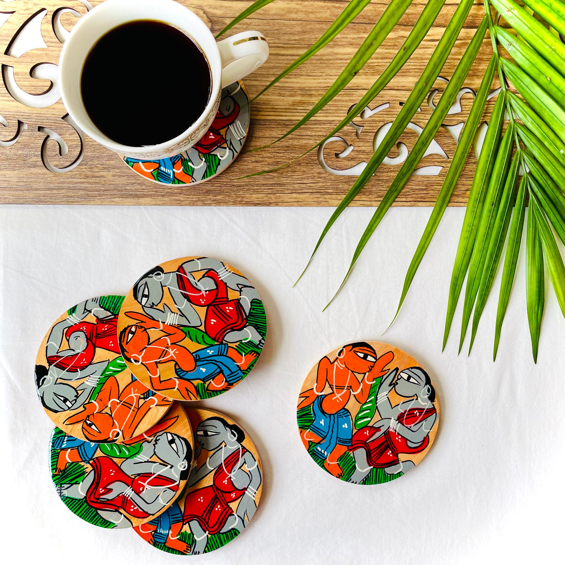 Five pure mango wood round wood coasters hand painted with tribal characters dancing are placed near a teacup filled with black tea with leaves in the background