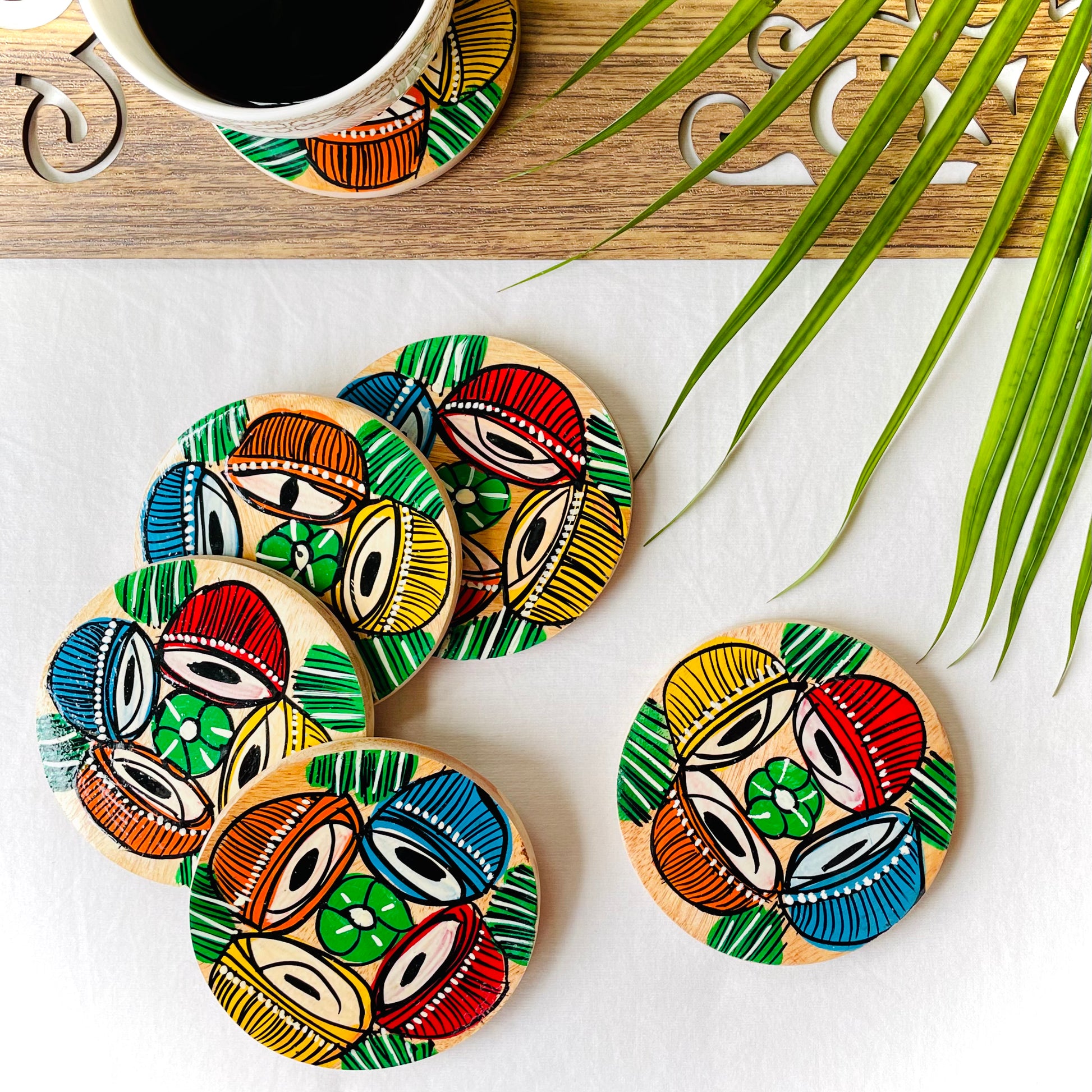 Five pure mango wood round wood coasters painted with four tablas, a type of Indian classical musical instrument in each wood coaster are placed near a teacup filled with black tea with leaves in the background
