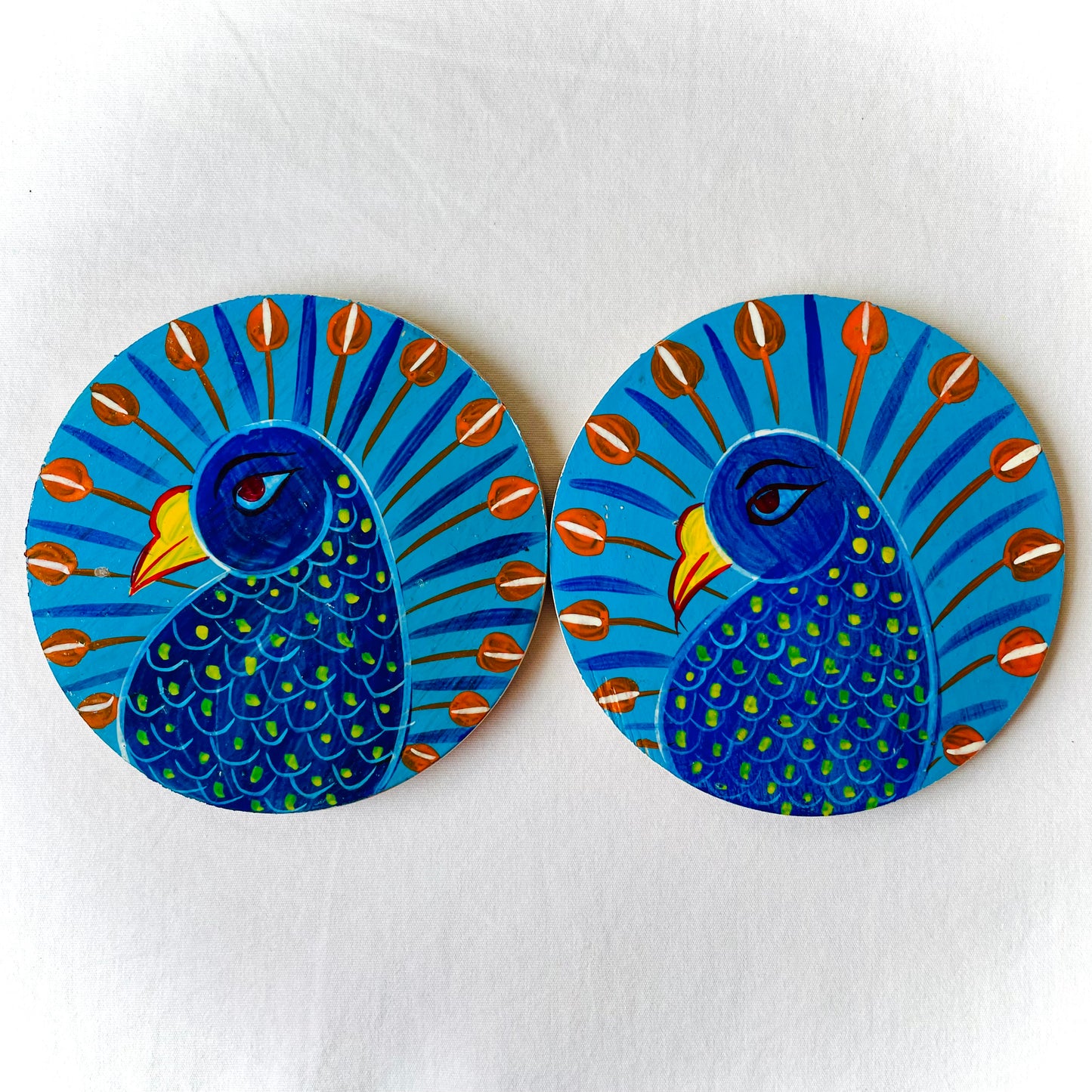 Two round wood coasters displayed against a white background, feature two peacocks with blue bodies, red feathers and yellow beaks