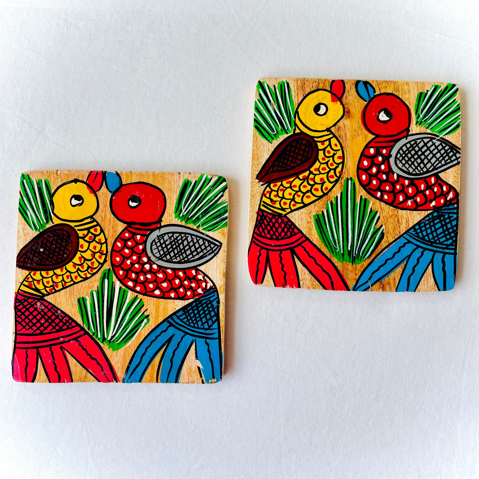 Two square wood coasters displayed against a white background, feature one yellow bird with red feathers and a beak and one red with blue feathers and a beak along with some leaves in each wood coaster