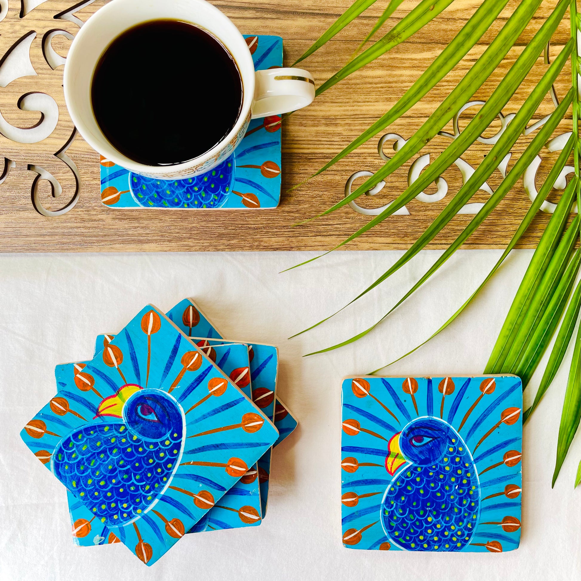 Five pure mango wood square wood coasters painted with peacocks having blue bodies, red feathers and yellow beaks are placed near a teacup on a wood coaster filled with black tea with leaves in the background