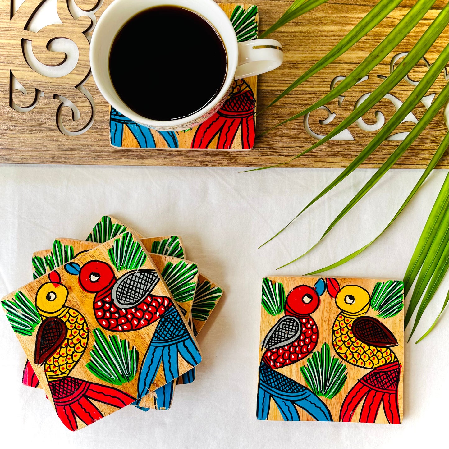 Five pure mango wood square wood coasters painted with one yellow bird with red feathers and a beak and one red with blue feathers and a beak along with some leaf in each wood coaster are placed near a teacup on a wood coaster filled with black tea with leaves in the background