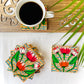 Five pure mango wood square wood coasters painted with four lotus with leaves and water in each wood coaster are placed near a teacup on a wood coaster filled with black tea with leaves in the background