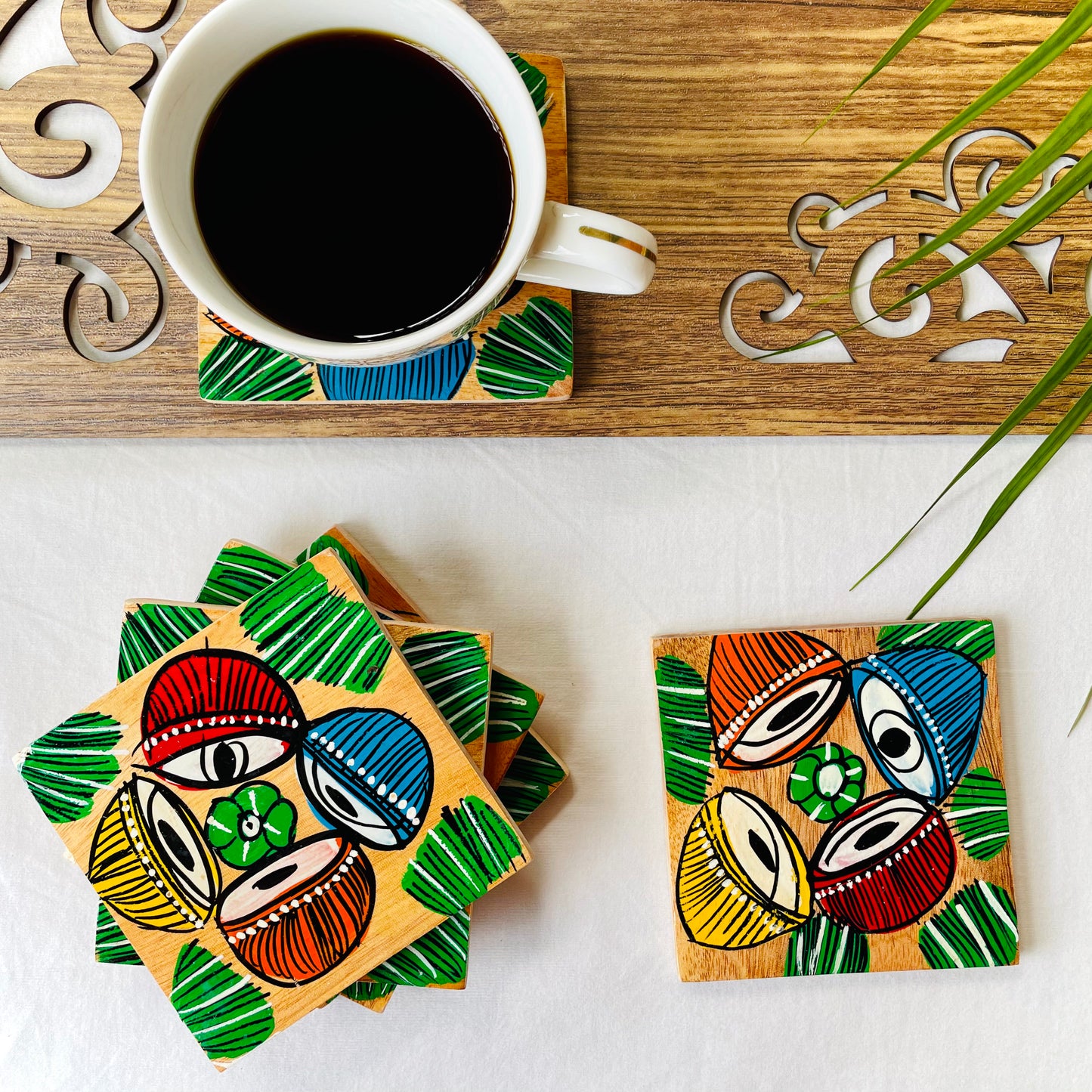 Five pure mango wood square wood coasters painted with four tablas, a type of Indian classical musical instrument in each wood coaster are placed near a teacup on a wood coaster filled with black tea with leaves in the background