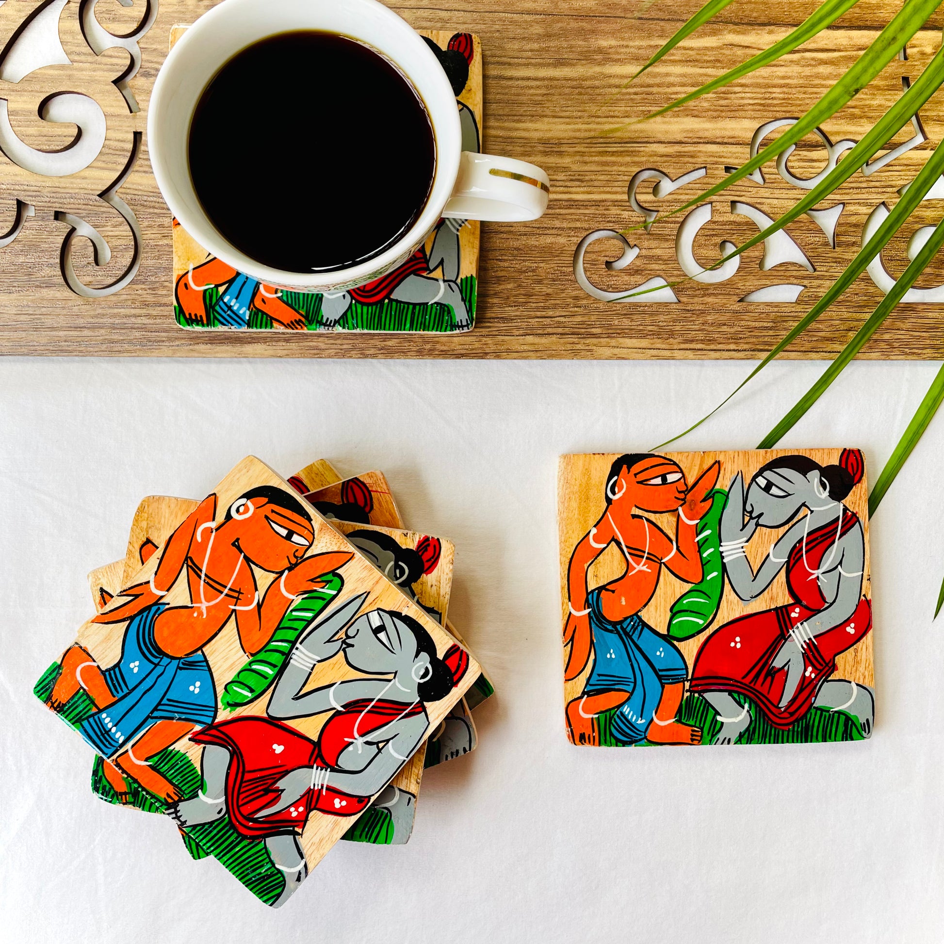 Five pure mango wood square wood coasters painted with tribal characters dancing are placed near a teacup on top of a wood coaster filled with black tea with leaves in the background