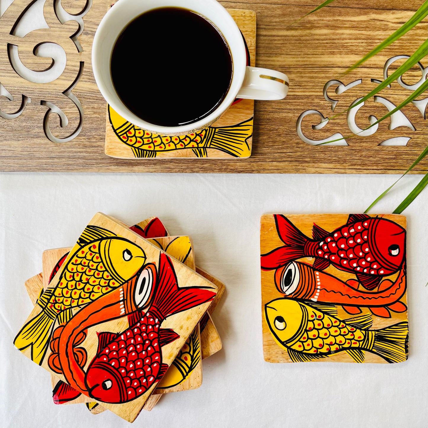 Five pure mango wood square wood coasters painted with one yellow fish and one redfish along with an Indian classical musical instrument in each wood coaster are placed near a teacup on a wood coaster filled with black tea with leaves in the background