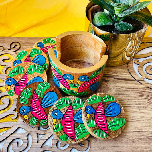 An empty coaster holder placed in front of a plant pot along with 6 round wooden coasters, all hand painted with bird and leaf motifs.