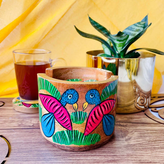 pure mango wood round wooden coaster holder with two birds painted on it, is being displayed with a warm cup of black tea and a plant pot in the background.