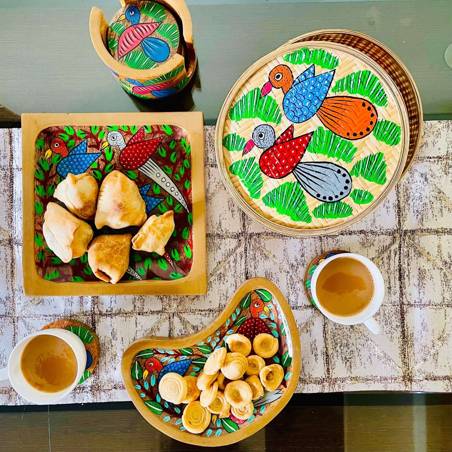 One moon-shaped pure mango wooden platter filled with snacks, one pure mango wood square wood serving tray filled with samosas arranged on a dining table along with a round wood coaster set, bamboo box, and tea cups; all having birds painted on it