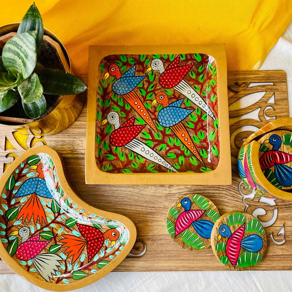 One pure mango Wood square Wood serving tray/trinket tray hand-painted with two blue birds and two white birds surrounded by tree branches, one moon-shaped pure mango wood serving tray/trinket tray hand-painted with three birds and pure mango wood round wood coaster set of 6 hand-painted with blue birds placed on a brown wood board beside a plant pot.