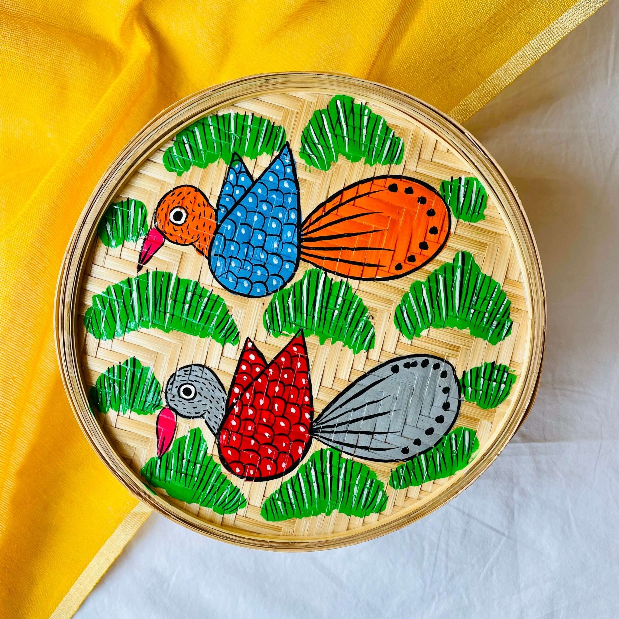 A handwoven bamboo box hand painted with one orange bird having blue wings and one grey bird having red wings flying over a green field is displayed against a yellow and white background