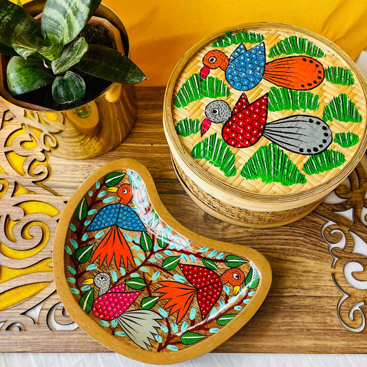 One pure mango wood moon-shaped wood serving tray/trinket tray hand-painted with three birds surrounded by tree branches, a one-hand woven bamboo fruit box, placed on a brown wood board beside a plant pot.