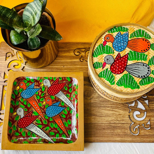 One pure mango wood square wood serving tray/trinket tray hand painted with four birds surrounded by tree branches, a one-hand woven bamboo fruit box hand painted with two birds flying over a field, placed on a brown wood board beside a plant pot.