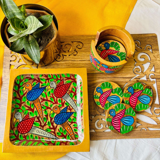 One pure mango wood square wood serving tray/trinket tray hand painted with four birds surrounded by tree branches, 3 round pure mango wood coster hand painted with blue coloured bird motifs are placed on a brown wood board beside a plant pot and round wood coaster holder.