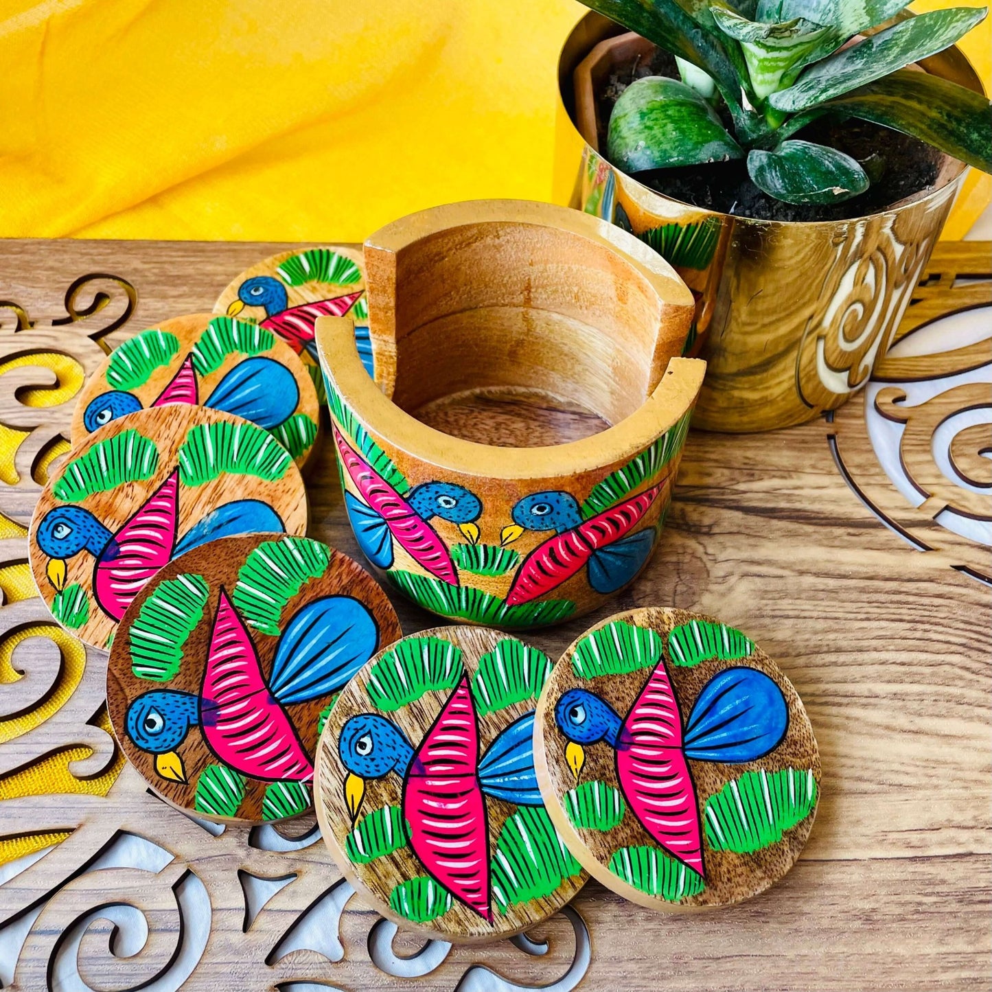 Six pure mango wood round coaster sets of 6 hand painted with blue coloured birds having pink wings arranged on a brown wood board beside a round wood coaster holder and plant pot.