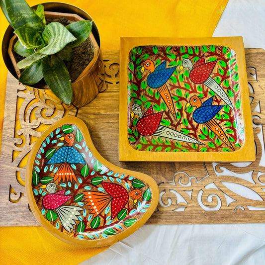One pure mango Wood square Wood serving tray/trinket tray hand-painted with two blue birds and two white birds surrounded by tree branches and one moon-shaped pure mango wood serving tray/trinket tray are placed on a brown wood board beside a plant pot.
