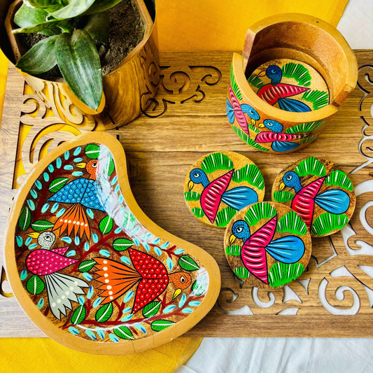 One pure mango wood, moon-shaped wood serving tray/trinket tray hand-painted with three birds surrounded by tree branches, 3 round pure mango wood coster hand painted with blue coloured bird motifs are placed on a brown wood board beside a plant pot and round wood coaster holder.