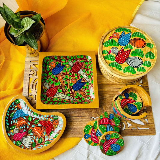 One pure mango Wood square Wood serving tray/trinket tray hand-painted with two blue birds and two white birds surrounded by tree branches, one moon-shaped pure mango wood serving tray/trinket tray hand-painted with three birds, one handwoven bamboo fruit box with a motif of birds flying over a field and pure mango wood round wood coaster set of 6 hand-painted with blue birds placed on a brown wood board beside a plant pot.