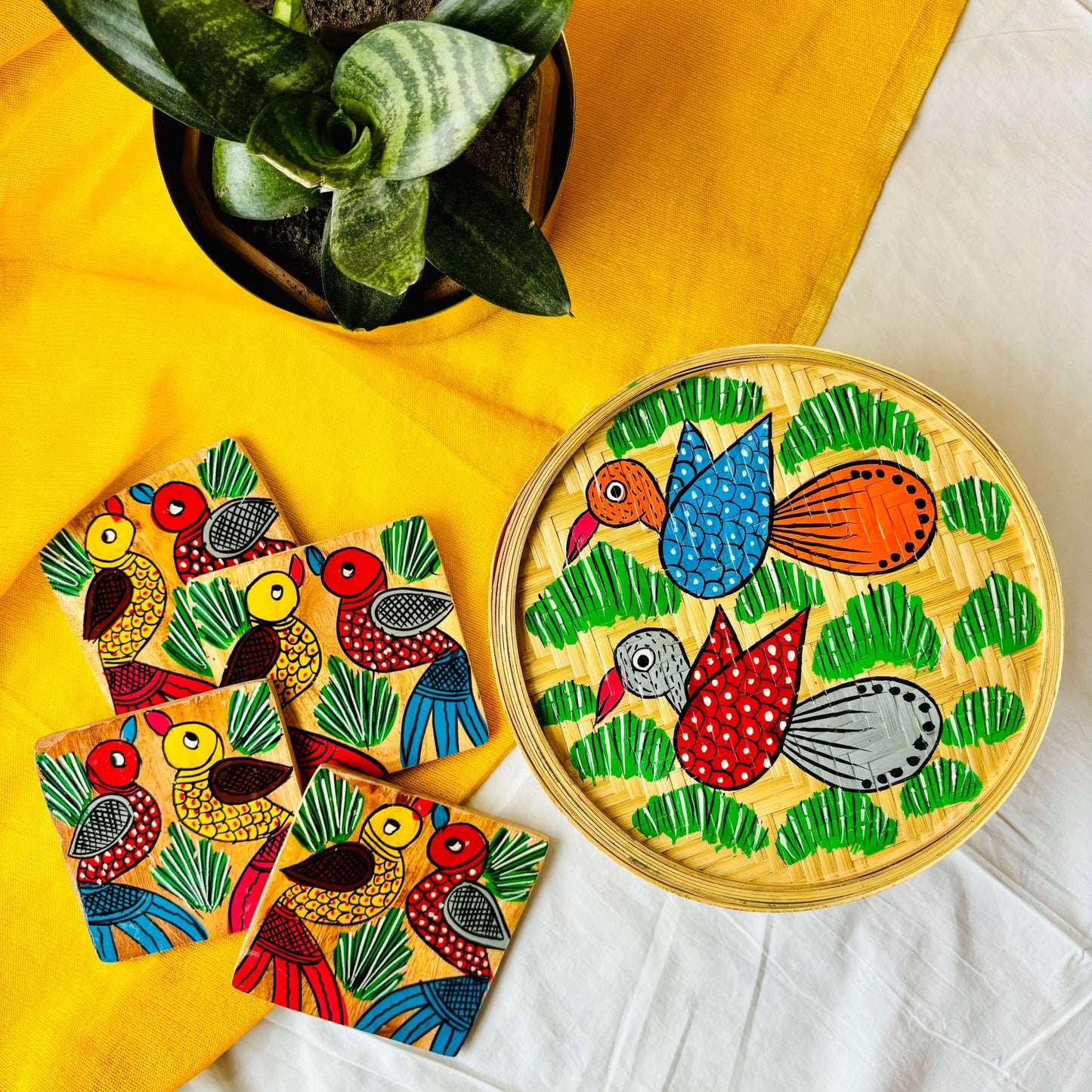 One handwoven bamboo fruit box with a motif of two birds flying over a field and a set of four square wood coasters with a motif of one yellow and one red bird are displaced against a yellow background with a plant pot beside them
