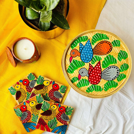One handwoven bamboo fruit box with a motif of two birds flying over a field, a set of four square wood coasters with a motif of one yellow and one red bird and a double wick scented candle displaced against a yellow background with a plant pot beside them